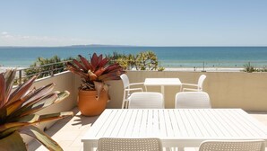 Une terrasse avec une table et des chaises blanches, un pot de fleurs et une vue sur la plage et l’océan.