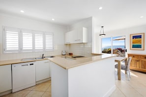 A modern kitchen with white cabinetry, a large island, and a view of the sea through a sliding door. - Portofino 7 - Beachfront Penthouse (Noosa Heads)