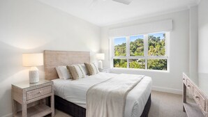 Une chambre à coucher comprenant un lit, des lampes de chevet, une table de chevet, une fenêtre donnant sur un paysage verdoyant et un ventilateur de plafond.