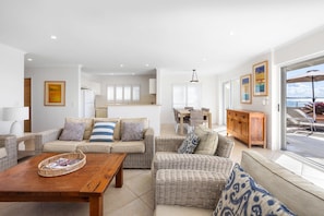A spacious living area with a sofa set, a wooden coffee table, and a dining table with chairs. There is a kitchen visible in the background, and a balcony with outdoor furniture. - Portofino 7 - Beachfront Penthouse (Noosa Heads)