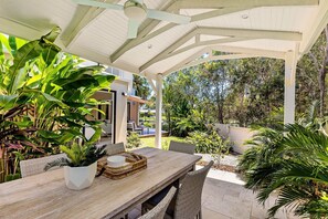 An outdoor patio with a wooden table and chairs, surrounded by lush greenery and a covered structure. - Quay Largo - Riverside Haven (Noosaville)