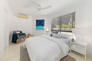 A bedroom with a bed, a chair, a bedside table, a lamp, a ceiling fan, and a window with blinds. - Quay Largo - Riverside Haven (Noosaville)