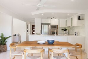 A modern kitchen with white cabinetry, a wooden dining table, and a ceiling fan. - Quay Largo: Riverside Haven (Noosaville)