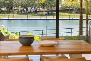 A wooden table with two bowls, overlooking a calm water body with a fence and trees in the background. - Quay Largo - Riverside Haven (Noosaville)