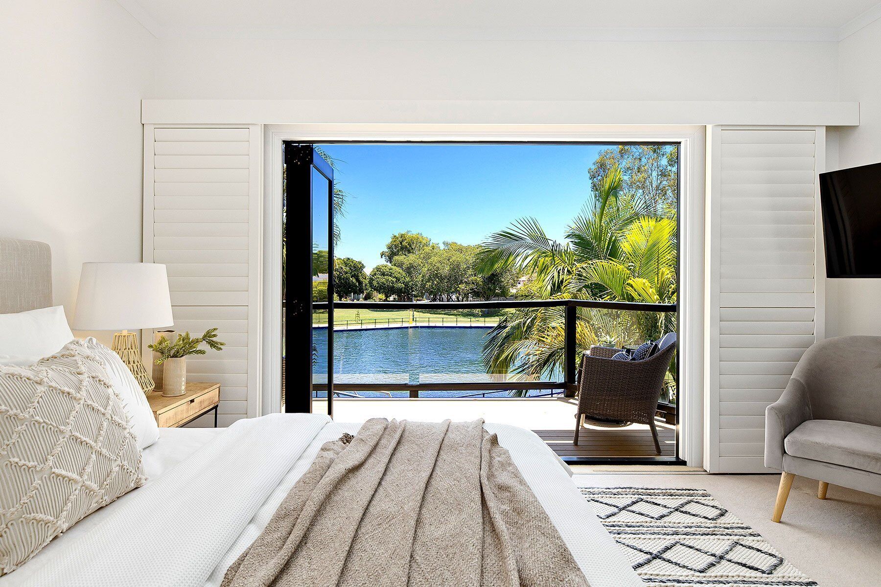 A bedroom with a bed, a nightstand, a lamp, a chair, and a view of a water body and greenery through an open door.