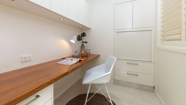 A compact kitchen with white cabinetry, a wooden countertop, a white chair, and a window with blinds.