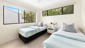 A bedroom with two beds, a nightstand, a lamp, and a window with a view of palm trees. - Soundhaven 1 - Prime Location (Noosa Heads)