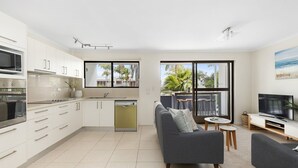 A modern kitchen with white cabinets, a dishwasher, and a dining area with a grey sofa and a small round table. - Soundhaven 1 - Prime Location (Noosa Heads)