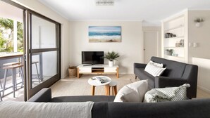 A modern living room with a large window, a flat-screen TV, a grey sofa, and a painting of a beach scene. - Soundhaven 1 - Prime Location (Noosa Heads)