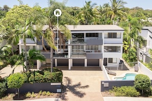 Un immeuble résidentiel moderne à deux étages, doté d’une piscine et entouré d’une végétation luxuriante et de palmiers.