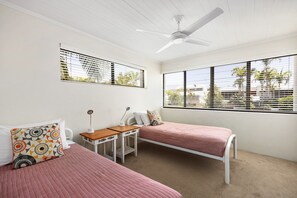 A bedroom with two beds, a desk, a chair, a ceiling fan, and large windows with blinds. - Soundhaven 2 - Steps to Hastings St (Noosa Parade)