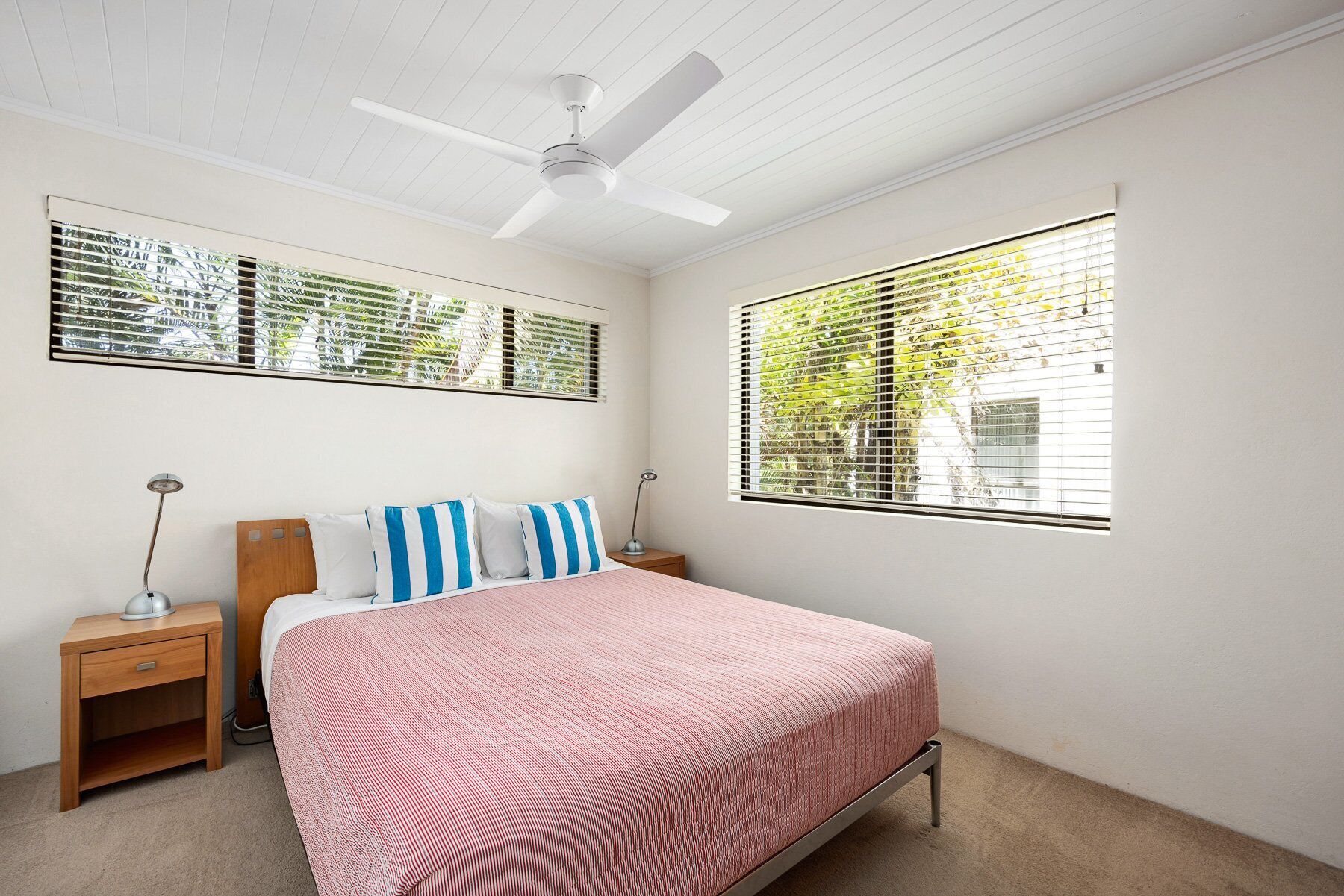 A bedroom with a bed, two bedside tables, a ceiling fan, and two windows with blinds.