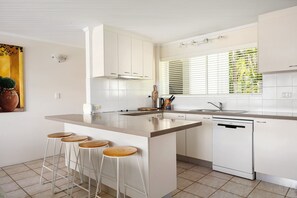 Une cuisine moderne dotée d’un îlot central, de tabourets de bar, d’un lave-vaisselle et d’une fenêtre avec des stores.