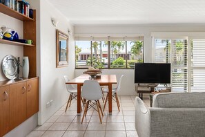 Un salon moderne comprenant un coin repas, un téléviseur et offrant une vue sur les palmiers à travers de grandes fenêtres.