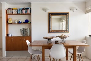 A dining area with a wooden table, white chairs, a mirror with a decorative frame, and a bookshelf with books and decorative items. - Soundhaven 2 - Steps to Hastings St (Noosa Parade)