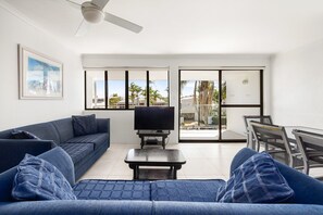 A modern living room with a sofa, a coffee table, and a TV. There is a balcony with a view of palm trees and buildings.