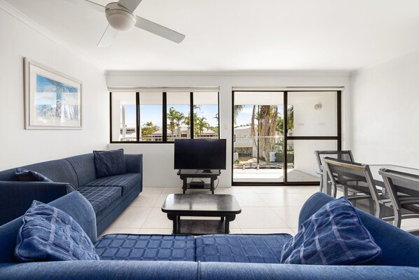 A modern living room with a sofa, a coffee table, and a TV. There is a balcony with a view of palm trees and buildings.