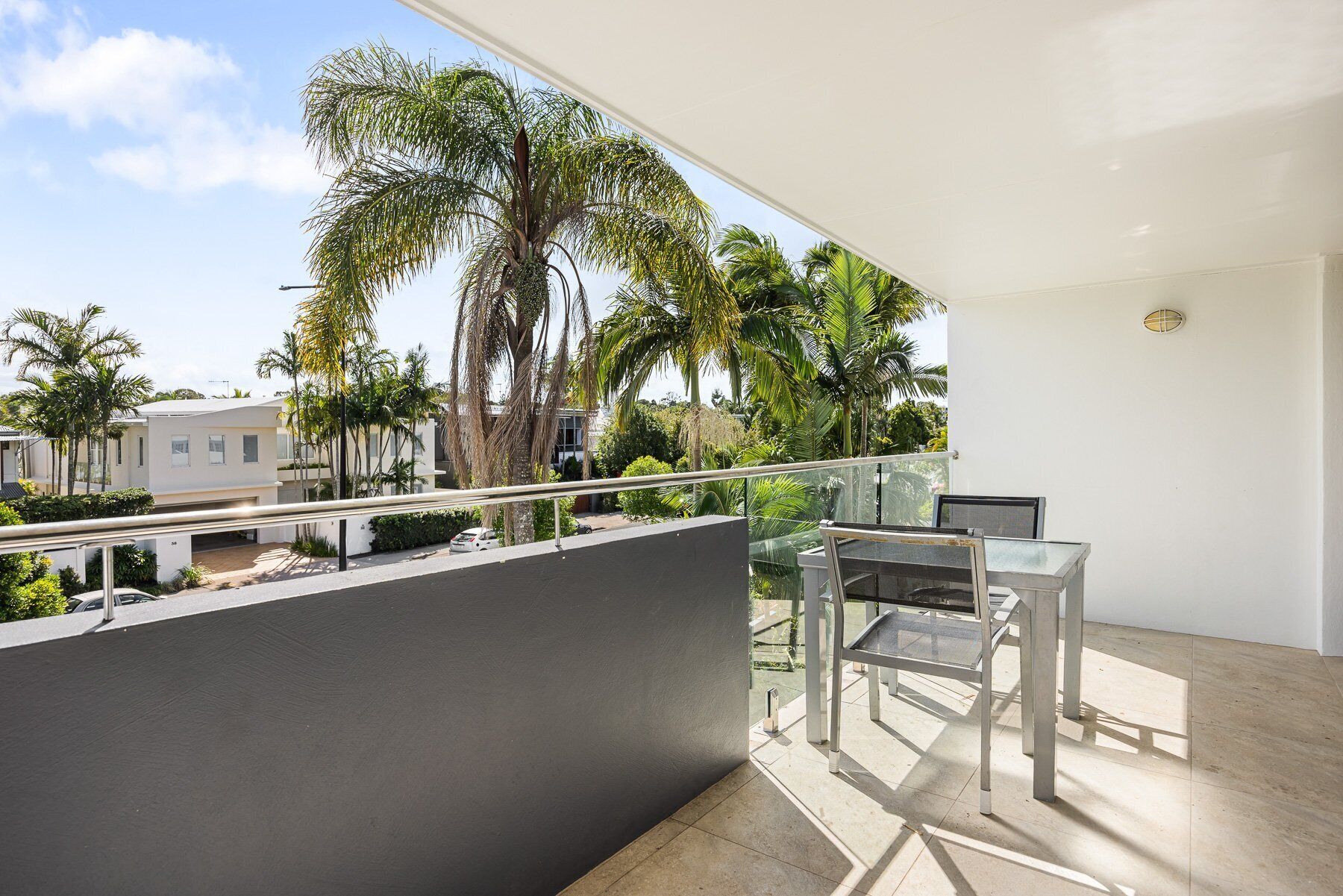 Un balcon doté d’une table et de chaises en verre, donnant sur un quartier résidentiel avec des palmiers et des maisons.