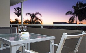 An outdoor dining area with a table and chairs, overlooking palm trees and a sunset. - Soundhaven 4 - Noosa Parade Value (Noosa Heads)