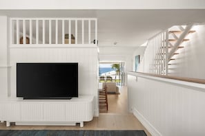 A modern interior with a flat-screen TV on a white entertainment unit, a staircase with wooden railings, and a view of the outdoors.