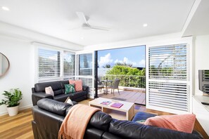 A modern living room with a black leather sofa, a coffee table, and a TV. There is a balcony with a white railing and a view of greenery.