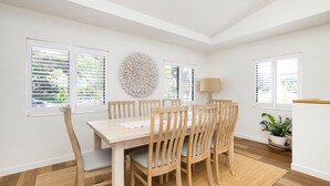 A dining area with a wooden table and chairs, a round wall decoration, and windows with blinds.