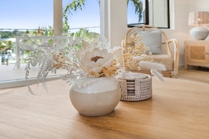 A room with a large window, a wicker side table, and a white vase with flowers.