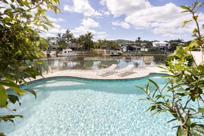 A clear swimming pool with lounge chairs, surrounded by palm trees and a dock with a boat. - St Tropez 12 - Haven with Water View & Pool (Noosa Heads)