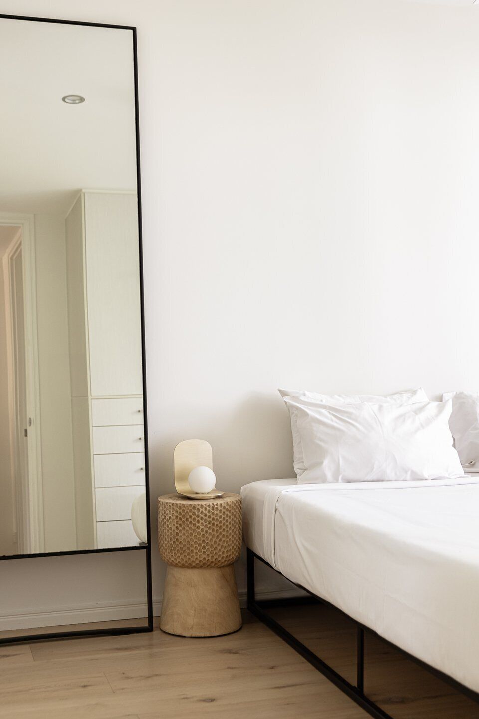 A bedroom with a bed, white bedding, a round stool, a mirror, and a wardrobe.