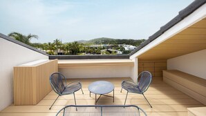 Terraço no telhado com vista para uma área residencial, duas cadeiras, uma mesa redonda pequena e um banco de madeira.