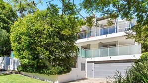 En modern bostadsbyggnad i två våningar med en balkong, ett garage och frodig grönska.