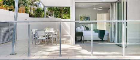 A modern outdoor pool area with a glass balustrade, a dining set, and a bedroom with a bed and a ceiling fan.