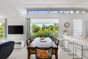A modern dining area with a long table, chairs, a TV, and a view of greenery through large windows.