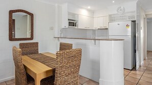 A compact kitchen with white cabinets, a microwave, a refrigerator, and a dining area with wicker chairs and a wooden table. - Villa Aqua - Riverside Retreat with Pool (Noosaville)