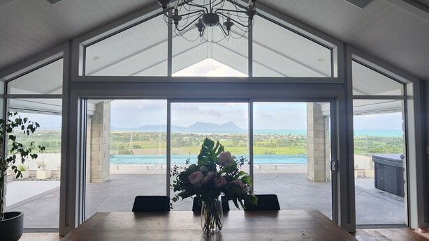 Un vaste espace dédié à la salle à manger, avec une grande table et des chaises, un lustre, et une vue sur une piscine et un paysage verdoyant grâce à de grandes fenêtres.
