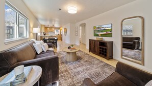 A modern living room with a brown leather sofa, a coffee table, and a TV. There is a kitchen in the background with wooden cabinets and a dining area. - BEST location in Bend! ~HOT TUB!~ (Bend)