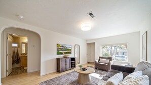 A modern living room with a sofa, a coffee table, and a TV. There is a kitchen area with cabinets and a window. - BEST location in Bend! ~HOT TUB!~ (Bend)