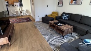 A modern living room with a grey sofa, a wooden coffee table, and a yellow armchair. There is a kitchen area in the background with wooden cabinets and a dining table.