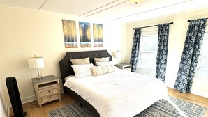 A bedroom with a large bed, a nightstand, a lamp, a ceiling fan, and a window with curtains.