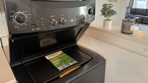 A front-loading washing machine with a control panel, a detergent packet, and a plant on the countertop.