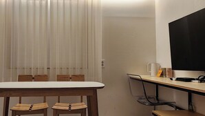 A small dining area with a table and chairs, a TV mounted on the wall, and a window with sheer curtains.