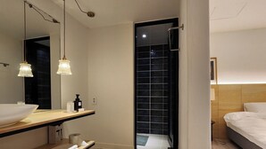 A modern hotel room with a wooden floor, a large mirror, a bedside table with toiletries, a shower area, and a bed.
