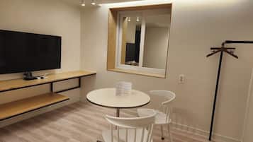Una habitación pequeña con una mesa redonda, dos sillas blancas, un televisor en un estante, un perchero y un espejo en la pared.
