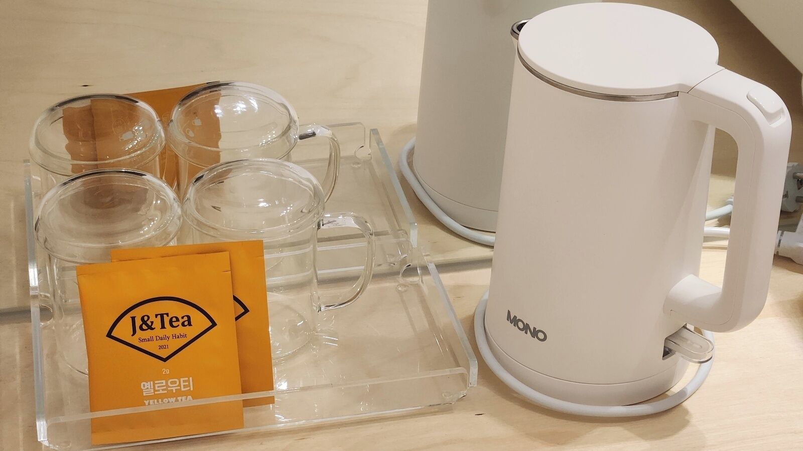 A clear glass mug with a handle, a white electric kettle, and a tray with four clear glass cups and a yellow tag labeled "J&Tea".
