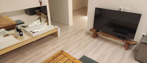 A modern, minimalist room with a wooden bench, a small white table with a sink, a flat-screen TV mounted on a wooden stand, and a refrigerator.