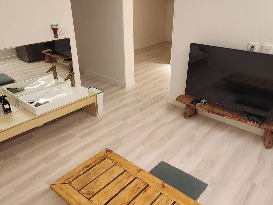 Una habitación moderna y minimalista con un banco de madera, una pequeña mesa blanca con un lavamanos, un televisor de pantalla plana montado en un atril de madera y un refrigerador.