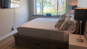 Un dormitorio con cama, ventilador de techo, ventana con vistas a árboles y playa, y l ámpara de noche.