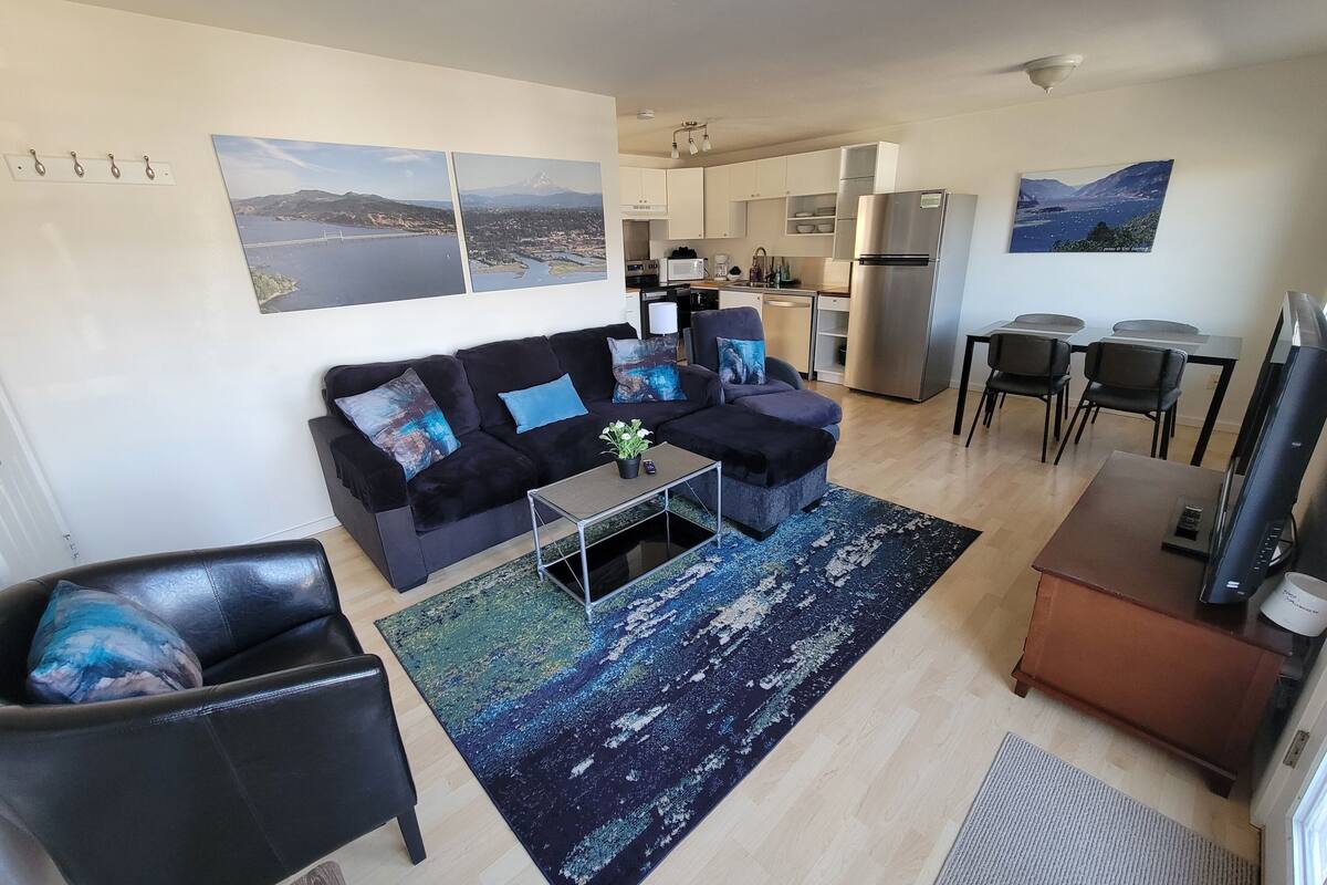 Una sala de estar moderna con un sofá negro, una mesa de centro y un televisor. Se observa una zona de cocina con un refrigerador y armarios.
