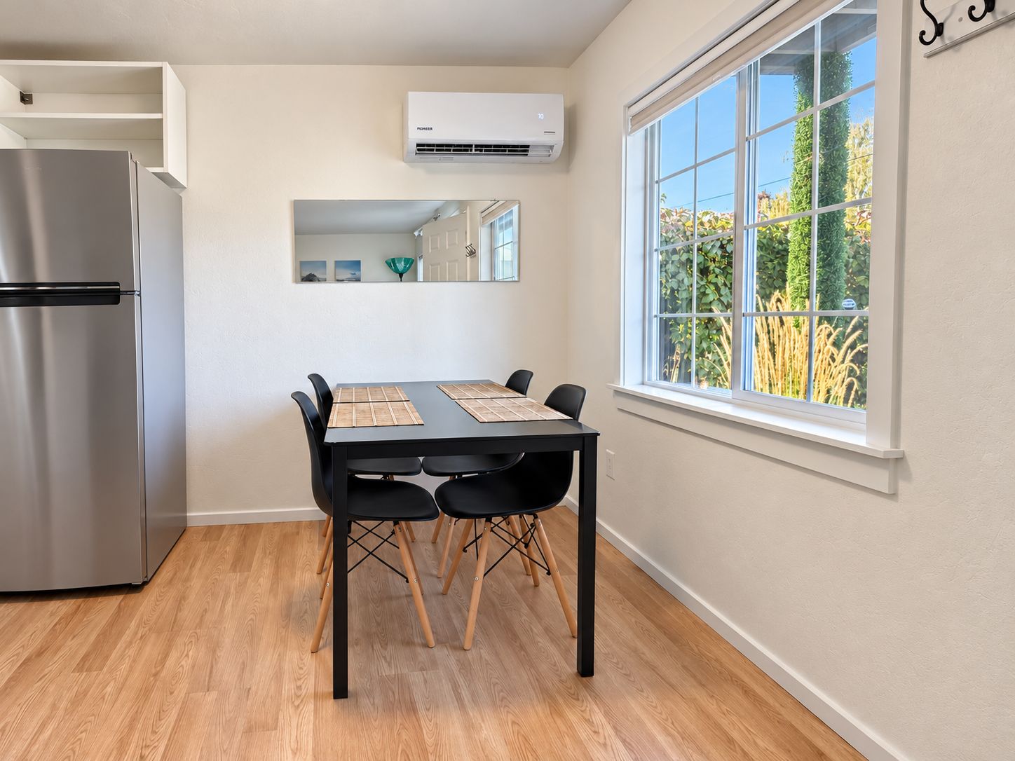 A modern kitchen with a stainless steel refrigerator, a dining table with four chairs, and a large window with a view of greenery.