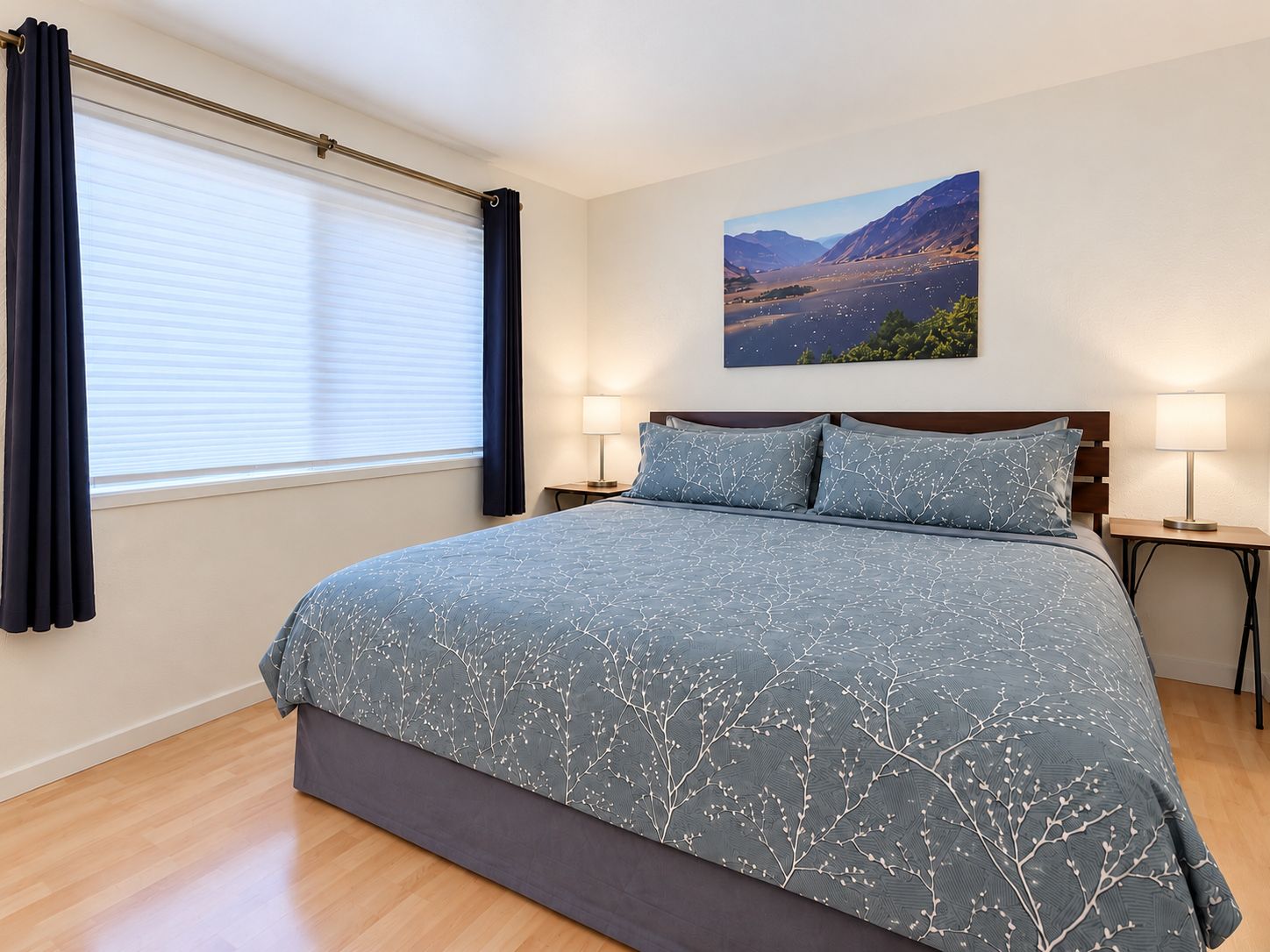 A bedroom with a large bed, two bedside tables with lamps, a window with curtains, and a landscape painting on the wall.